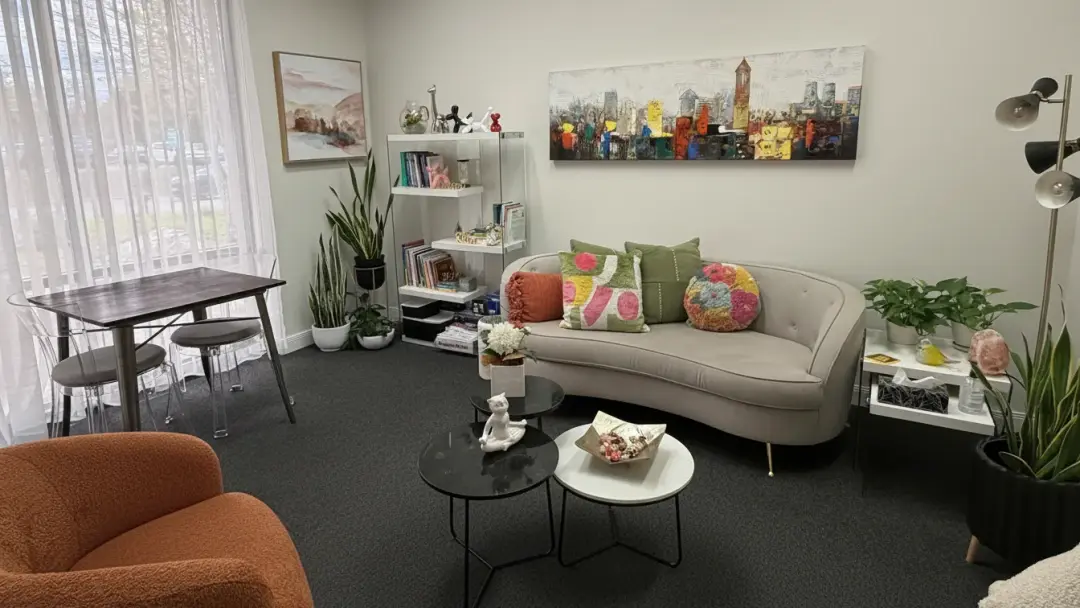 Ciepły gabinet psychologiczny w Oakville: kremowa sofa z kolorowymi poduszkami pod obrazem panoramy miasta, rośliny i okno z delikatną firaną